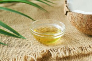 A close-up of coconut oil in a glass bowl on burlap, surrounded by coconut shell and leaves.